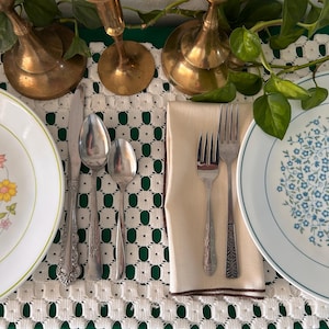 Könnte beinhalten: Eine Tischdekoration mit zwei Tellern, Besteck und einer gefalteten Serviette. Ein Teller hat ein Blumendesign mit gelben und orangen Blumen, der andere ein blaues Blumenmuster. Das Besteck umfasst ein Messer, Löffel und Gabeln. Zwei Messing-Kerzenhalter sind sichtbar.