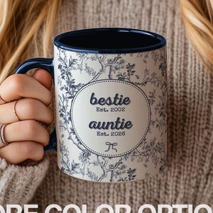 May include: A white ceramic mug with a blue floral pattern and a navy blue interior and handle. The mug has the words "bestie" and "auntie" in a circular design, with the text "Est. 2002" and "Est. 2026". The words "MORE COLOR OPTIONS" are at the bottom.