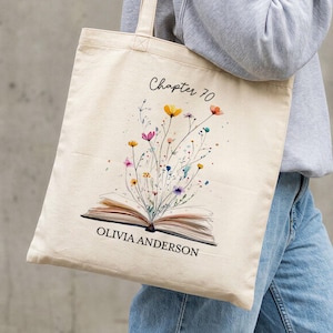 May include: A beige canvas tote bag featuring a watercolor design of an open book with colorful flowers blooming from it. The text "Chapter 70" is above the design, and "OLIVIA ANDERSON" is below. The bag has long handles.