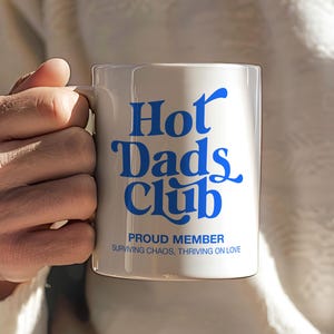 Peut inclure: Mug en céramique blanche avec l'inscription "Hot Dads Club" en bleu. En dessous, on peut lire "Proud Member" et la phrase "Surviving Chaos, Thriving on Love" en plus petit. La tasse est tenue par une personne portant un pull blanc.