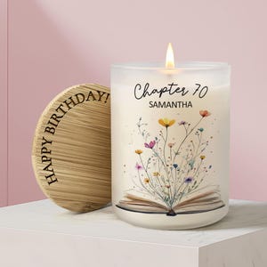 May include: A frosted glass candle with a lit wick, featuring a printed graphic of an open book with colorful wildflowers emerging from the pages. The text reads Chapter 70 SAMANTHA. A round bamboo lid with the text HAPPY BIRTHDAY is placed beside it.