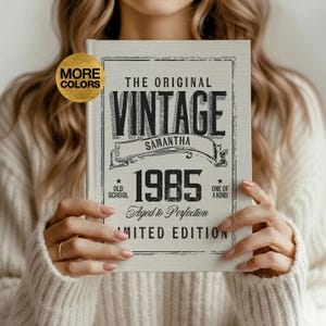 May include: A vintage-style book with the text "THE ORIGINAL VINTAGE SAMANTHA 1985 Aged to Perfection LIMITED EDITION." The book is held by a person wearing a white sweater. A gold circle with the words "MORE COLORS" is in the upper left corner.