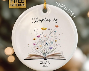May include: A round white ceramic ornament hanging from a gold string. The front features the text "Chapter 16" above an illustration of an open book with colorful wildflowers sprouting from the pages. Below the book is the text "OLIVIA 2026".