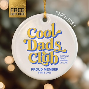 May include: A round white ceramic ornament with a gold hanging string. The text reads Cool Dads Club in large yellow and blue letters, with smaller blue text stating Surviving Chaos Thriving On Love and Proud Member Since 2025.