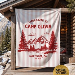 May include: A white and pink striped banner with the words "WELCOME TO CAMP OLIVIA" in red. Below the text is a red illustration of mountains, trees, and the text "LAKE TAHOE, CA EST. 2026". The banner is hanging on a wooden structure.