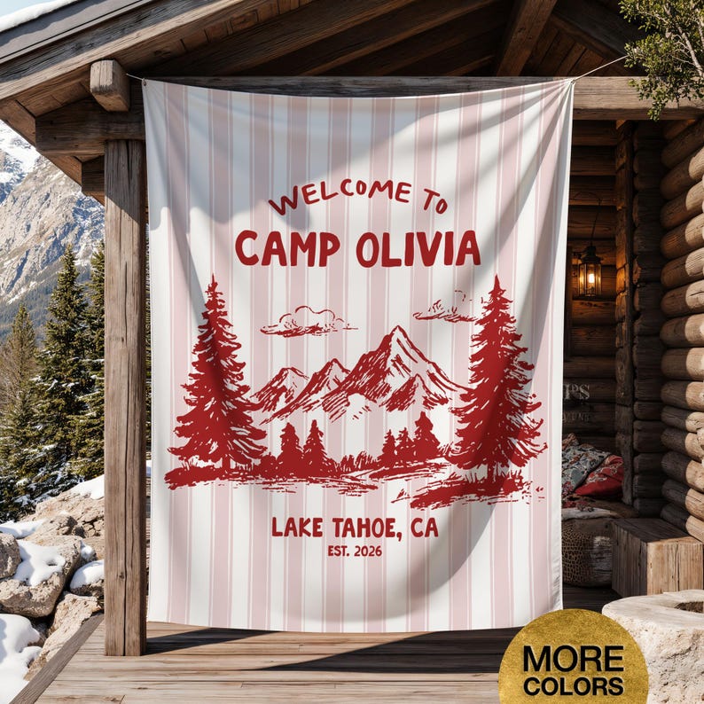 May include: A white and pink striped banner with the words "WELCOME TO CAMP OLIVIA" in red. Below the text is a red illustration of mountains, trees, and the text "LAKE TAHOE, CA EST. 2026". The banner is hanging on a wooden structure.