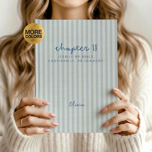 May include: A hardcover book with a light blue and white vertical striped cover. The text reads "chapter 18 LEGALLY AN ADULT. EMOTIONALLY, NO COMMENT." and the name "Olivia" is printed below. A gold circular badge in the corner says "MORE COLORS."
