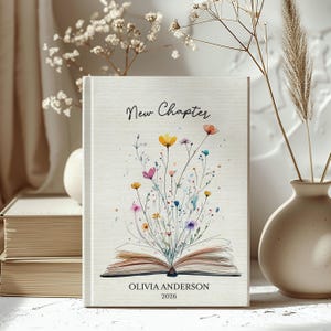 May include: A hardcover journal with the words "New Chapter" in script. The cover features a watercolor illustration of colorful wildflowers emerging from an open book. The name "Olivia Anderson" and the year "2026" are printed at the bottom.
