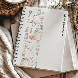 May include: A white spiral-bound notebook with a medical-themed watercolour design. The design includes a stethoscope, pills, syringes, and a nurse's hat. The name "OLIVIA ANDERSON RN" is printed vertically on the right side of the notebook.