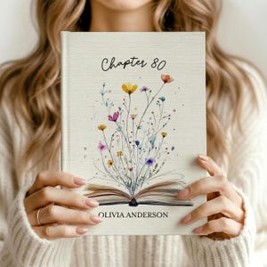 Puede incluir: Un libro de tapa dura con una cubierta texturizada de color crema que presenta una ilustración en acuarela de un libro abierto con flores silvestres de colores que brotan de las páginas. El texto dice Chapter 80 y OLIVIA ANDERSON.