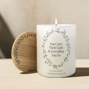 May include: A white candle with a floral wreath design and the text "You Carry Their Light In Everything You Do." The candle has a lit wick and a wooden lid with the words "Sending You Love."