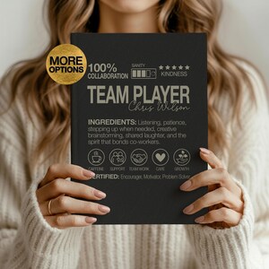 May include: A black canvas print with the words "TEAM PLAYER" and "Chris Wilson" in a gray font. The print includes the words "INGREDIENTS" and icons representing caffeine, support, teamwork, care, and growth.