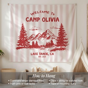 May include: A wall tapestry with a red and white striped background. The tapestry features a red illustration of mountains, trees, and the text "WELCOME TO CAMP OLIVIA LAKE TAHOE, CA EST. 2026". Instructions on how to hang the tapestry are also included.