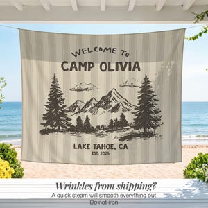 May include: A beige and white striped outdoor banner with the text "WELCOME TO CAMP OLIVIA" and "LAKE TAHOE, CA EST. 2026". The banner features a mountain and tree graphic. The banner is hanging on a porch overlooking a beach.