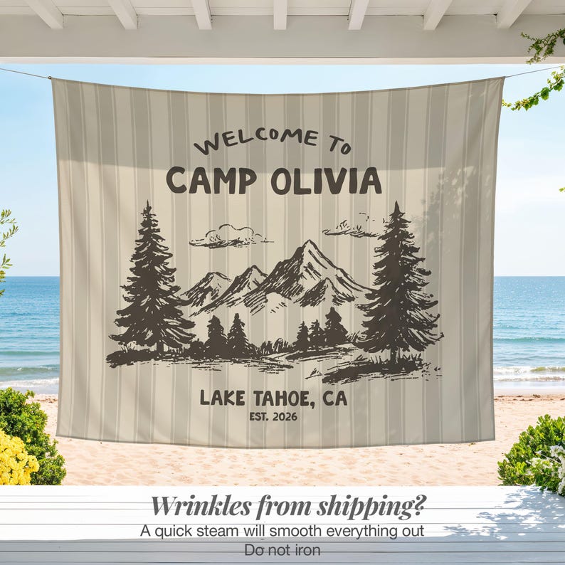May include: A beige and white striped outdoor banner with the text "WELCOME TO CAMP OLIVIA" and "LAKE TAHOE, CA EST. 2026". The banner features a mountain and tree graphic. The banner is hanging on a porch overlooking a beach.
