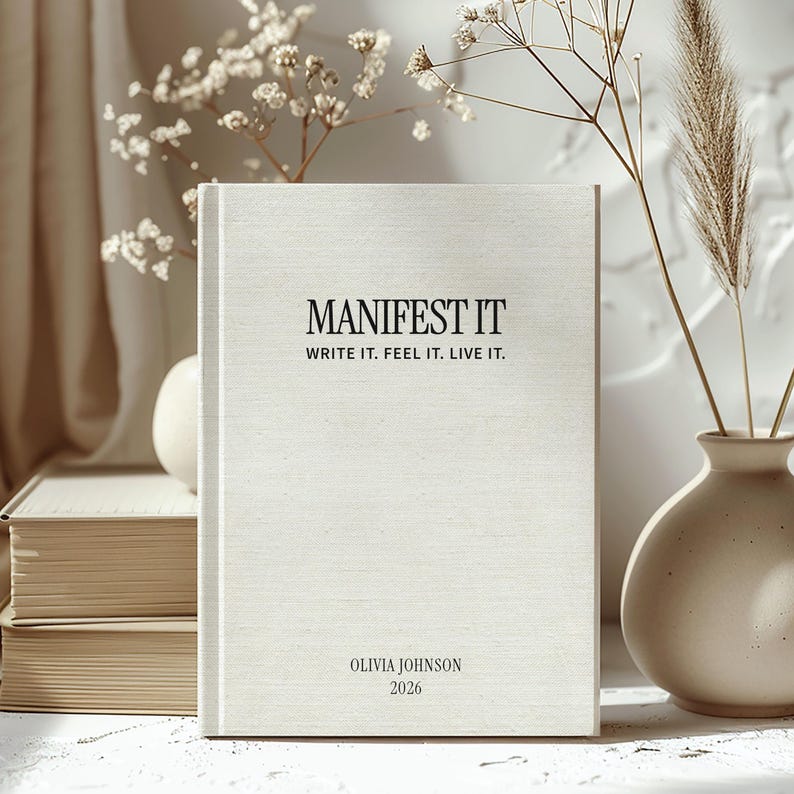 May include: A cream-colored journal with the words "MANIFEST IT" in black, along with "WRITE IT. FEEL IT. LIVE IT." The journal also includes the name "OLIVIA JOHNSON" and the year "2026".