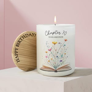 May include: A white candle with a floral design and the text "Chapter 70 Olivia Anderson." A wooden lid with "Happy Birthday!" is next to the candle. The candle is lit with a flame.