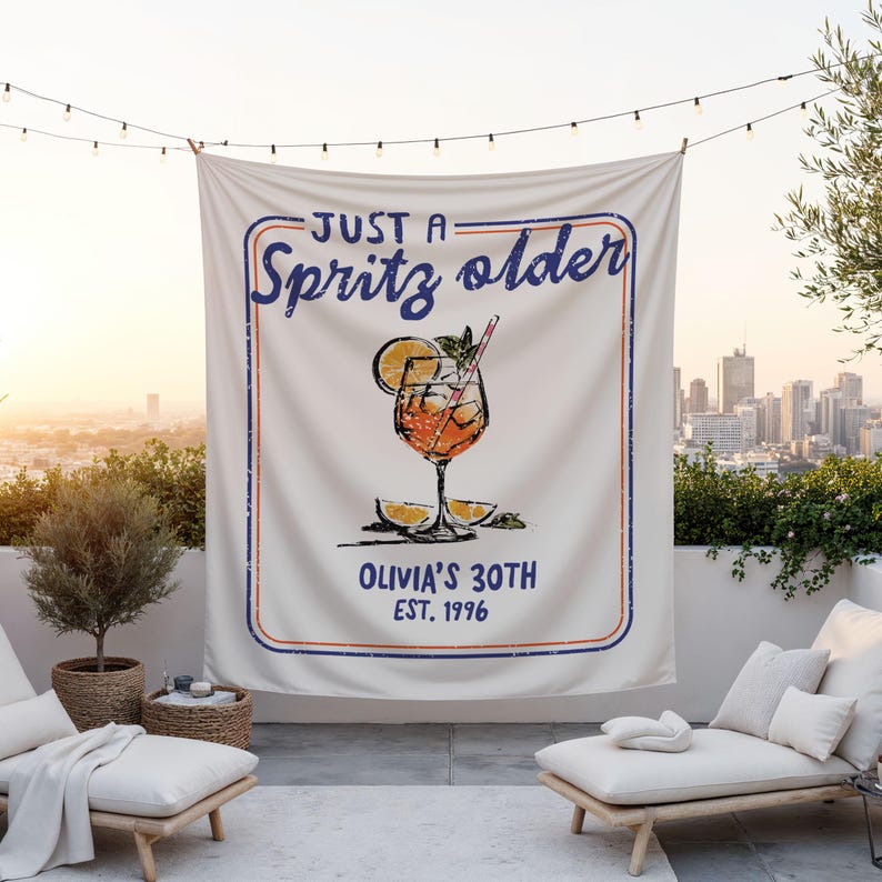 May include: A white tapestry featuring the text "JUST A Spritz older" in blue, with a cocktail graphic. Below the graphic is "OLIVIA'S 30TH EST. 1996". The tapestry is displayed outdoors with a city skyline visible in the background.