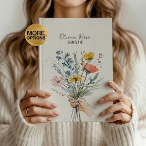 Pode incluir: Um diário de capa dura com uma ilustração de um buquê de flores. A capa apresenta o texto "Olivia Rose Chapter 18". O diário é segurado por uma pessoa vestindo um suéter creme. Um círculo dourado com as palavras "More Options" está no canto superior esquerdo.