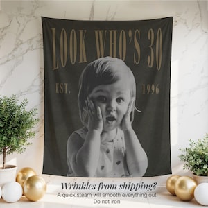 May include: A dark gray rectangular fabric wall hanging featuring a black and white photograph of a child with hands on their cheeks. The text "LOOK WHO'S 30" appears in large gold letters at the top, with "EST. 1996" printed in gold below the text.