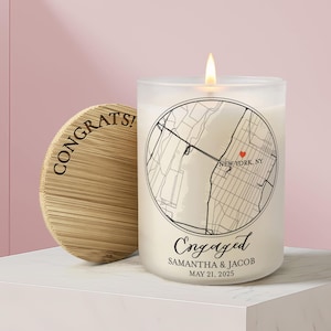 May include: A frosted glass candle with a lit wick, featuring a circular black map graphic of New York, NY with a red heart, the text Engaged, Samantha & Jacob, and May 21, 2025. Beside it is a round bamboo lid engraved with the word CONGRATS.