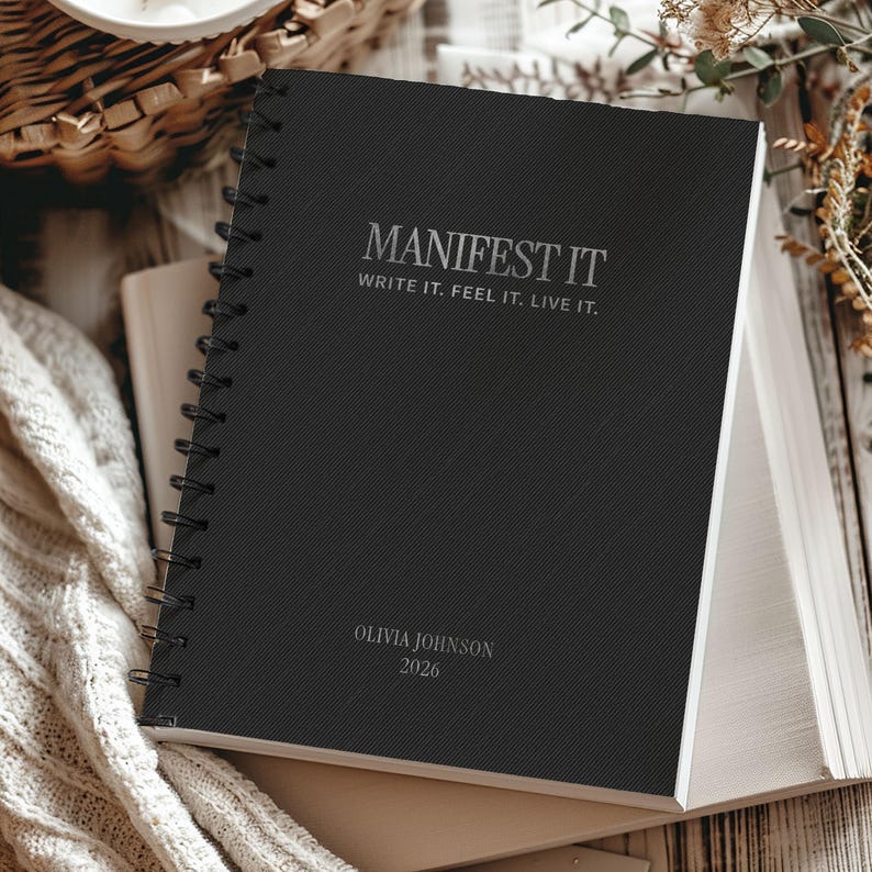 May include: Black spiral-bound notebook with the text "MANIFEST IT" and the tagline "WRITE IT. FEEL IT. LIVE IT." The notebook also includes the name "OLIVIA JOHNSON" and the year "2026". The notebook is resting on top of a book and a cream-colored knitted blanket.