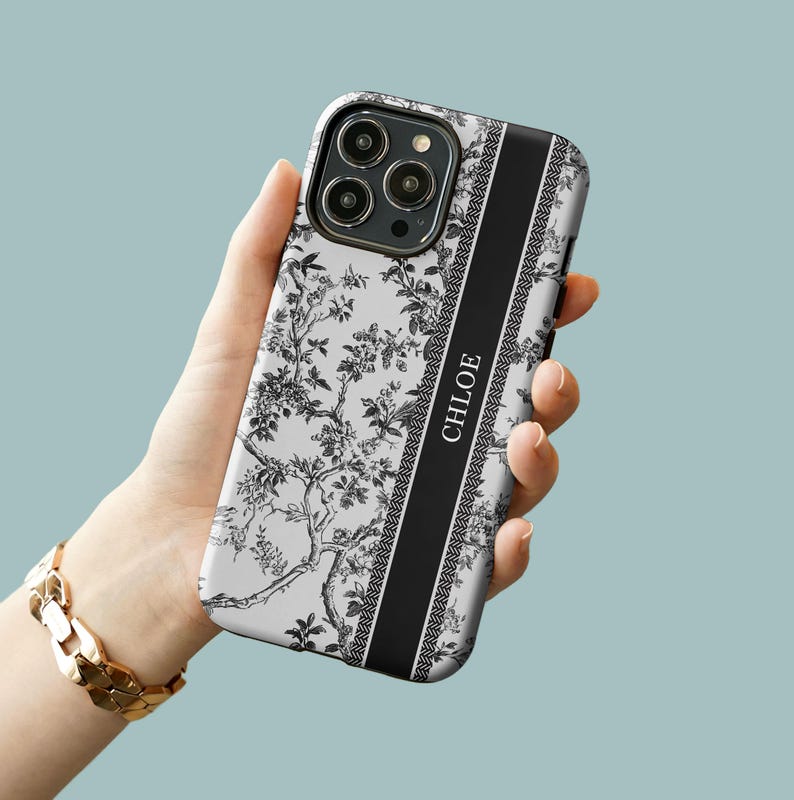 May include: A white phone case with a black and white floral pattern and a black stripe with the name "CHLOE" printed on it.