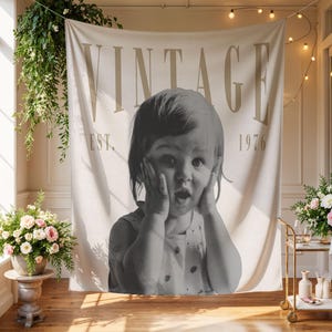 May include: A rectangular fabric wall tapestry featuring a large black and white photograph of a child with hands on their cheeks. The word VINTAGE is printed in large beige letters at the top, with EST. 1976 printed in smaller beige text below.