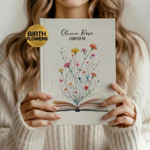 May include: A hardcover journal with the text "Olivia Rose Chapter 40" and a floral illustration. The journal cover features a gold "Birth Flowers" emblem. The journal is open, revealing the pages.