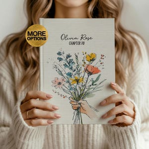 Puede incluir: Un diario de tapa dura con una ilustración de un ramo de flores. La portada presenta el texto "Olivia Rose Capítulo 30". El ramo incluye flores rosas, amarillas y naranjas con follaje verde y azul. El diario es sostenido por una persona que lleva un suéter color crema.