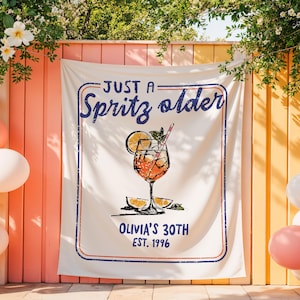 May include: A white banner with a blue and red border, featuring the text "JUST A Spritz older" and "OLIVIA'S 30TH EST. 1996." A graphic of a cocktail with an orange slice and a straw is in the center. The banner is hanging outdoors.