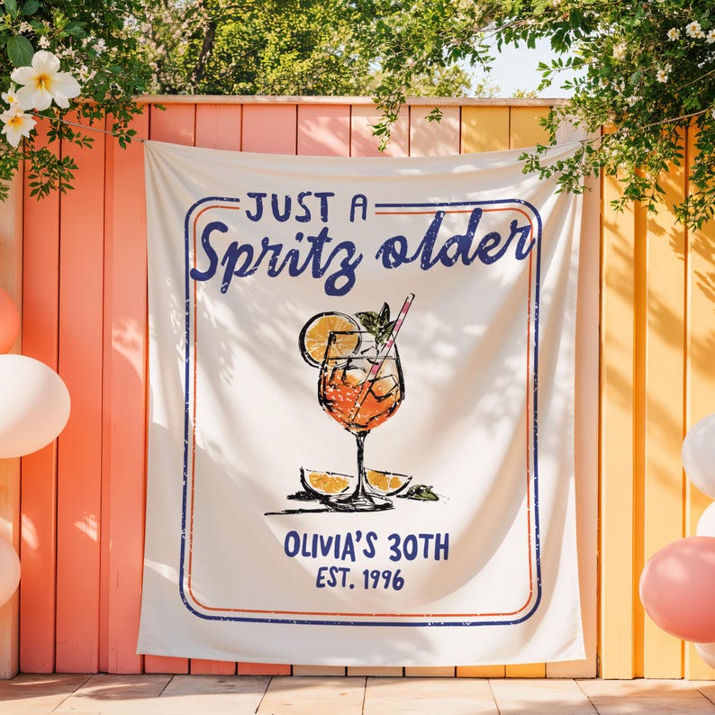 May include: A white banner with a blue and red border, featuring the text "JUST A Spritz older" and "OLIVIA'S 30TH EST. 1996." A graphic of a cocktail with an orange slice and a straw is in the center. The banner is hanging outdoors.