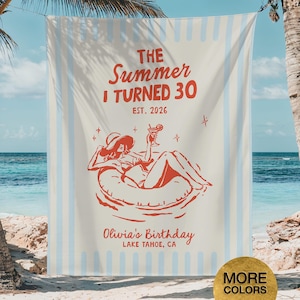 May include: A beach towel with a cream background and light blue vertical stripes. The towel features red text that reads "The Summer I Turned 30" and "Olivia's Birthday, Lake Tahoe, CA." A red illustration depicts a person relaxing on a float.