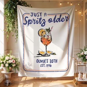 May include: White fabric backdrop with a blue and red border, featuring the text "Just a Spritz older" and a graphic of a cocktail with orange slices. Below the graphic is "Olivia's 30th Est. 1996."