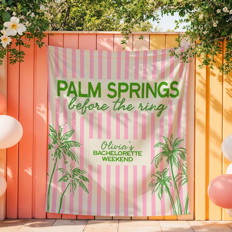 May include: A banner with the text "PALM SPRINGS before the ring" in green, with pink and white stripes. The banner also includes the text "Olivia's BACHELORETTE WEEKEND" and palm tree illustrations. The banner is hanging on a pink and yellow wall.