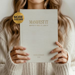 May include: A light beige journal with the words "MANIFEST IT" and "WRITE IT. FEEL IT. LIVE IT." in gold lettering. The journal also includes the name "OLIVIA JOHNSON" and the year "2026". A gold-colored circle with the words "MORE COLORS" is in the upper left corner.