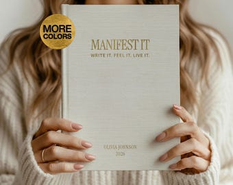 May include: A light beige journal with the words "MANIFEST IT" and "WRITE IT. FEEL IT. LIVE IT." in gold lettering. The journal also includes the name "OLIVIA JOHNSON" and the year "2026". A gold-colored circle with the words "MORE COLORS" is in the upper left corner.