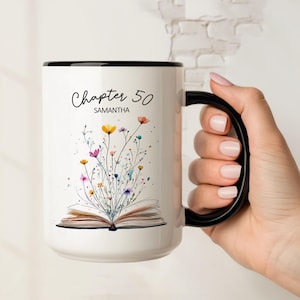 Puede incluir: Taza de cerámica blanca con asa y borde negros. La taza presenta el texto "Chapter 50" sobre el nombre "Samantha" y una ilustración de acuarela de flores coloridas que emergen de un libro abierto.