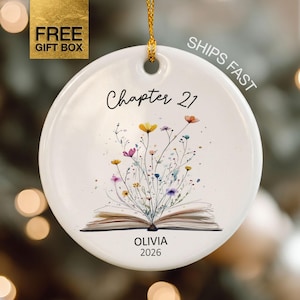 May include: A round white ceramic ornament hanging from a gold string. The front features an illustration of an open book with colorful wildflowers sprouting from the pages. The text reads Chapter 21, OLIVIA, and 2026 in black lettering.