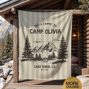 May include: A beige vertical striped banner with the text "WELCOME TO CAMP OLIVIA" and a mountain and tree graphic. Below the graphic is "LAKE TAHOE, CA EST. 2026". The banner is hanging on a wooden porch.