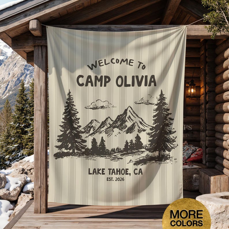 May include: A beige vertical striped banner with the text "WELCOME TO CAMP OLIVIA" and a mountain and tree graphic. Below the graphic is "LAKE TAHOE, CA EST. 2026". The banner is hanging on a wooden porch.
