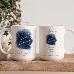 Op de afbeelding: Twee witte keramische mokken met een donkerblauw aquarelontwerp van een nachtelijke hemel met het sterrenbeeld Leeuw. De ene mok toont de naam Olivia en Leo, de andere de tekst Leo, Radiant, Generous, Confident en Main Character Energy.