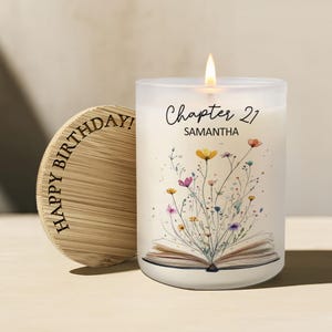 May include: A frosted glass candle with a lit wick, featuring a printed illustration of an open book with colorful wildflowers emerging from the pages. The text reads Chapter 21 and SAMANTHA. A round bamboo lid with HAPPY BIRTHDAY engraved is adjacent.