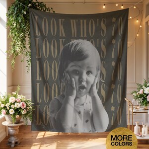 May include: A rectangular wall tapestry featuring a black and white photograph of a child with hands on their cheeks. The background is a dark gray fabric printed with the repeating gold text "LOOK WHO'S 70" in a vertical, stacked layout.
