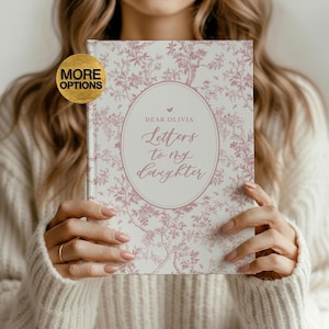 May include: A journal titled "Letters to My Daughter" is held. The cover features a pink floral pattern and an oval frame with the text "Dear Olivia". The journal is a hardcover book with a white background.