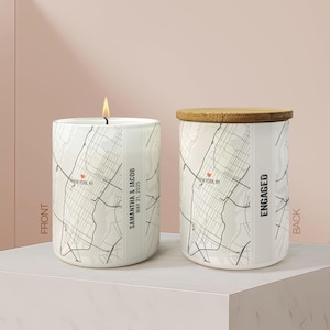 May include: Two white ceramic candles with a map design. One candle is lit, the other has a wooden lid. Text on the candles reads "New York, NY", "Samantha & Jacob May 21, 2025", and "Engaged". The candles are on a white surface.