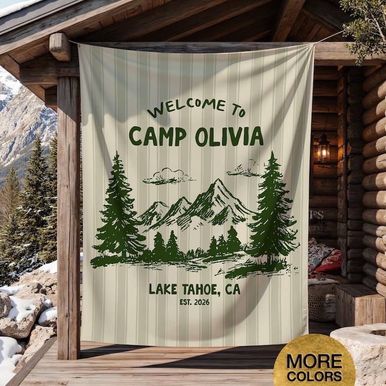 May include: A beige and white striped outdoor banner with the text "WELCOME TO CAMP OLIVIA" in green. Below the text is a green illustration of mountains, trees, and the text "LAKE TAHOE, CA EST. 2026". The banner is hanging on a wooden structure.