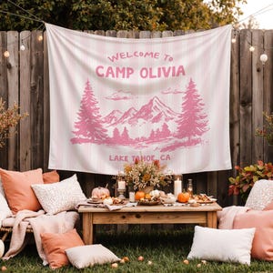 May include: A pink and white striped tapestry with the text "WELCOME TO CAMP OLIVIA" and a mountain and tree design. The tapestry is hanging on a wooden fence, with a table set for a gathering in front of it.