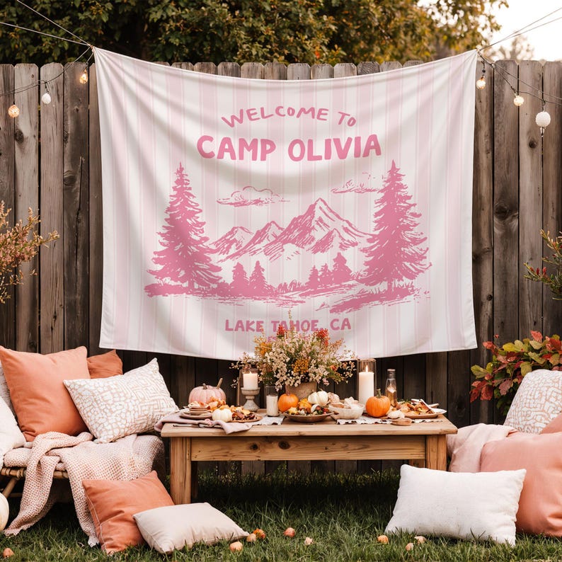 May include: A pink and white striped tapestry with the text "WELCOME TO CAMP OLIVIA" and a mountain and tree design. The tapestry is hanging on a wooden fence, with a table set for a gathering in front of it.