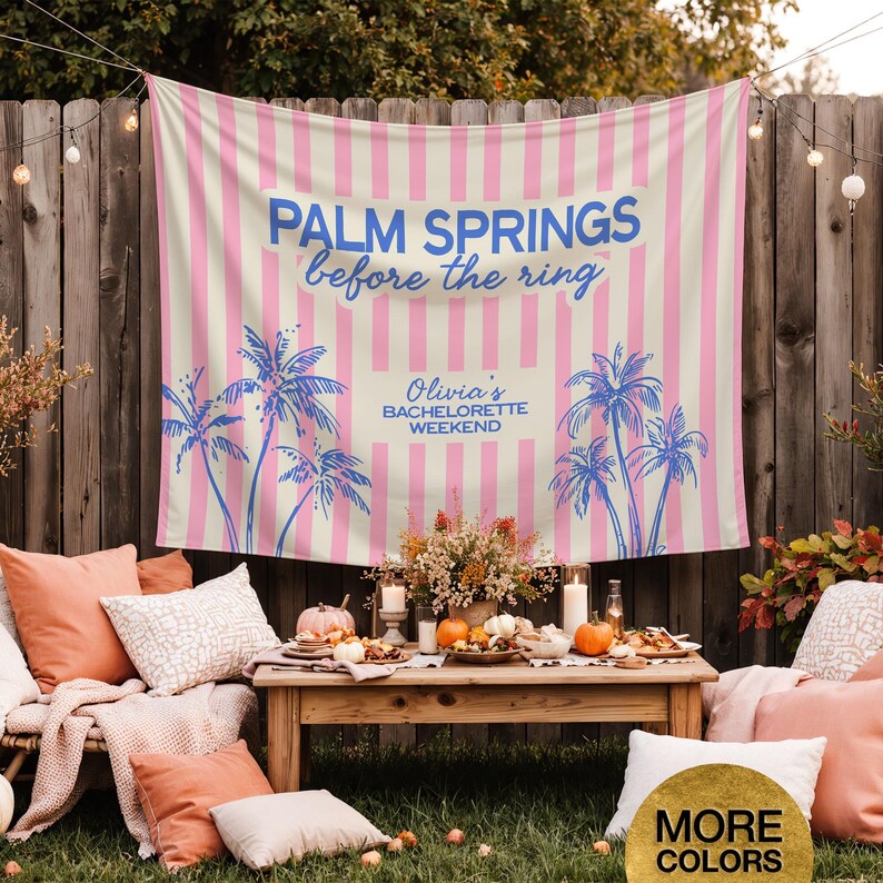 May include: A pink and cream striped backdrop with blue text that reads "Palm Springs before the ring" and "Olivia's Bachelorette Weekend". Palm tree illustrations are also featured. The scene includes a wooden table with food and pillows.